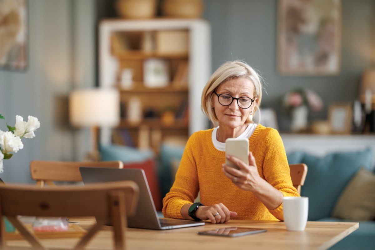 A woman at a kitchen table checking her phone beside a laptop