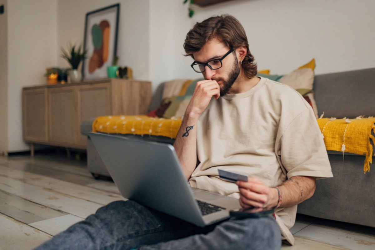 A man sitting on the floor with a laptop and a bank card, checking his account
